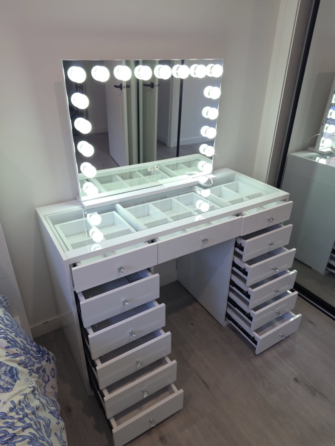 White glass-top vanity with open storage drawers and a lit Hollywood mirror beside a mirrored wardrobe.