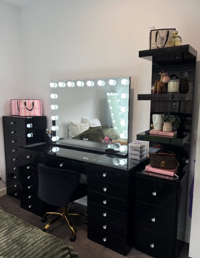 Black glass-top vanity with a lit Hollywood mirror, black swivel chair and matching storage drawers.