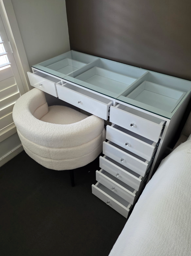 Compact white glass-top vanity with open drawers and a cream rounded chair beside window shutters.