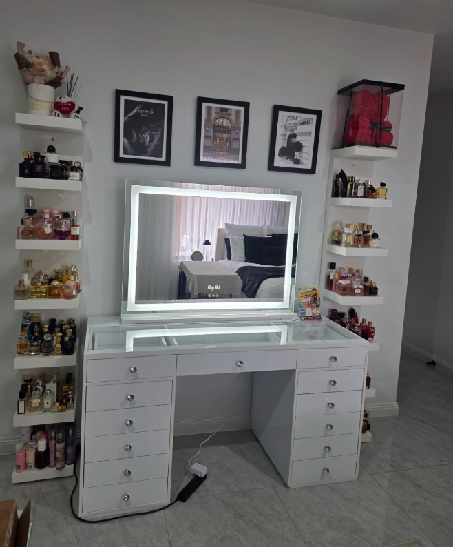 White glass-top vanity with an illuminated border mirror between shelves of perfume bottles.