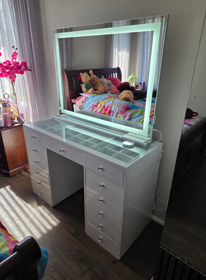 White glass-top vanity and illuminated border mirror beside a bedroom window with pink flowers.