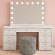 White makeup vanity with an illuminated Hollywood mirror, crystal-style drawer knobs and a pink tufted ottoman.