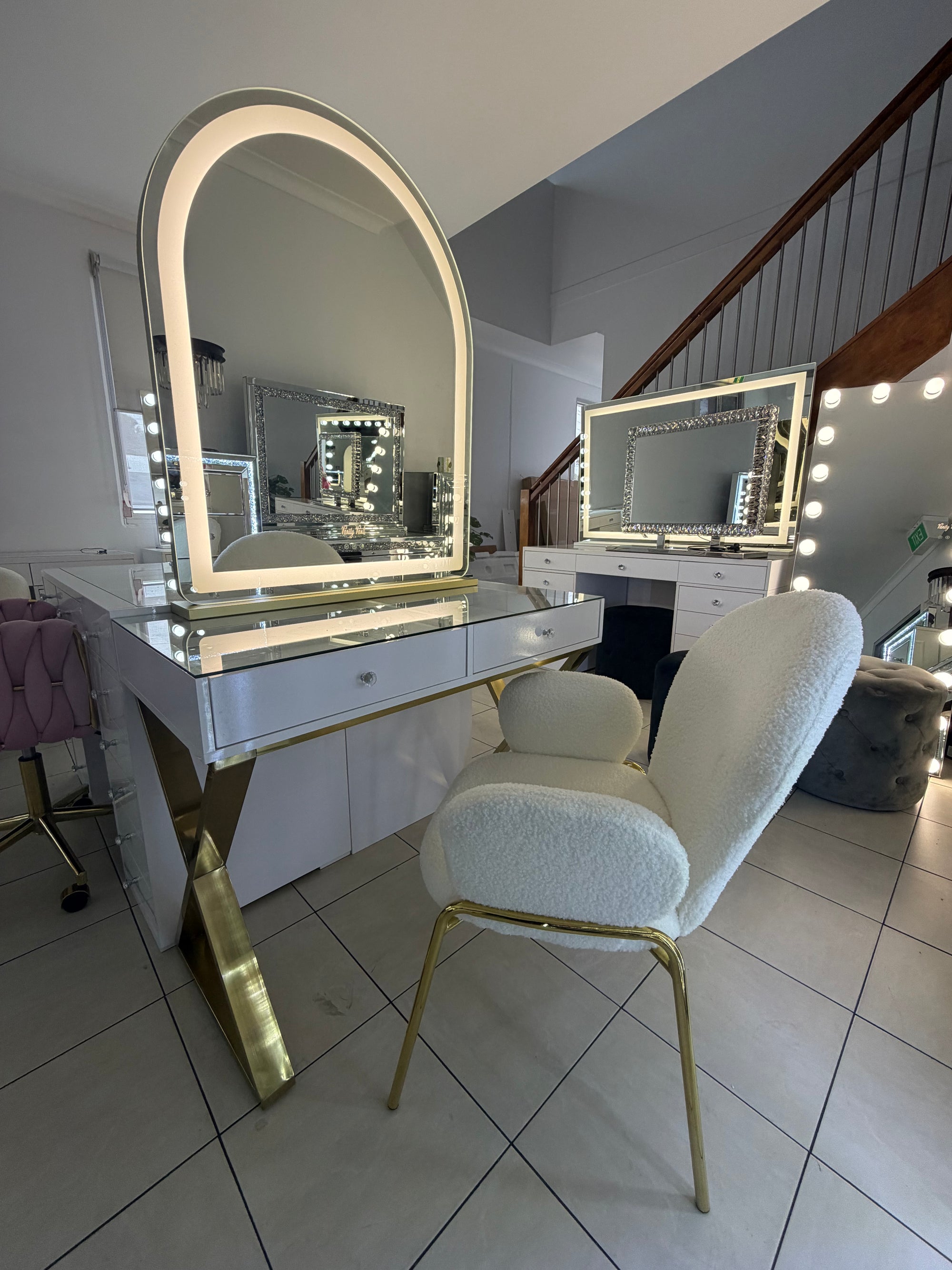A luxury vanity desk set featuring a large arched LED-backlit mirror, a mirrored drawer table with gold legs, and a plush cream bouclé armchair with gold metal legs, set in a modern home office.